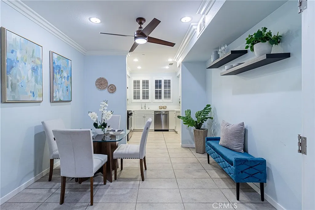 Dining area with round table, four chairs, ceiling fan, light blue walls, blue bench, wall art, and kitchen in background.