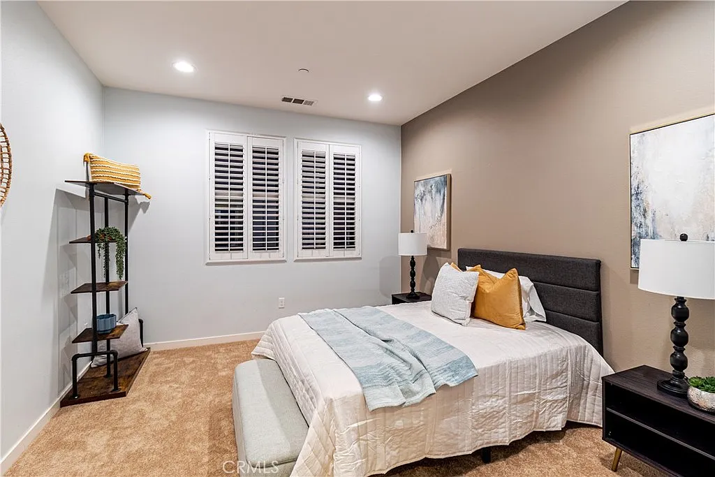 Bedroom with white and blue bed, yellow pillow, bedside lamps, window with shutters, painting, nightstand, shelf, and carpeted floor