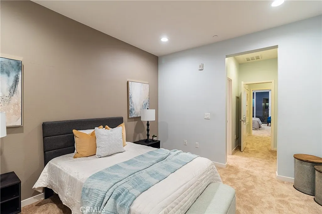 Bedroom with gray headboard bed, white bedding, decorative pillows, bedside lamps, neutral walls, doorway, and carpeted floor.