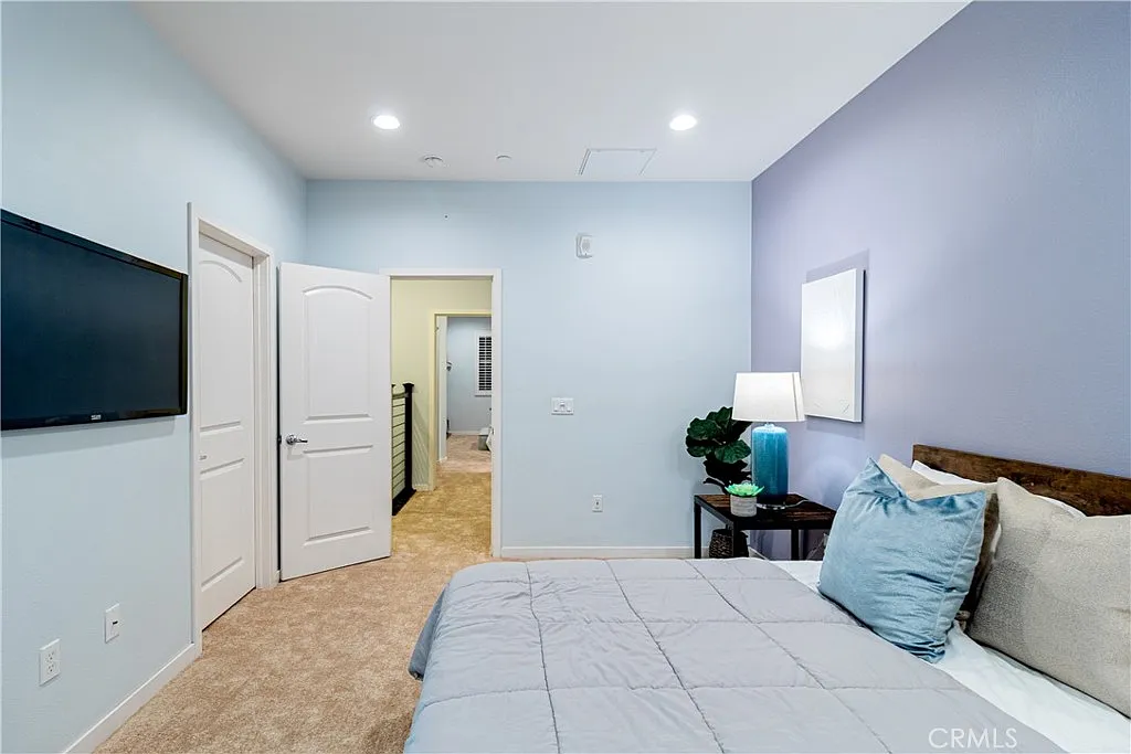 Blue bedroom with bed, pillows, nightstand with lamp, wall tv, two doors, and carpeted floor.