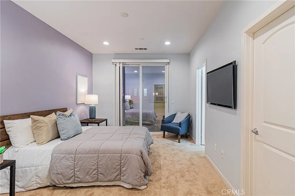 Bedroom with gray and beige bed, bedside lamps, blue chair, wall tv, and large windows.