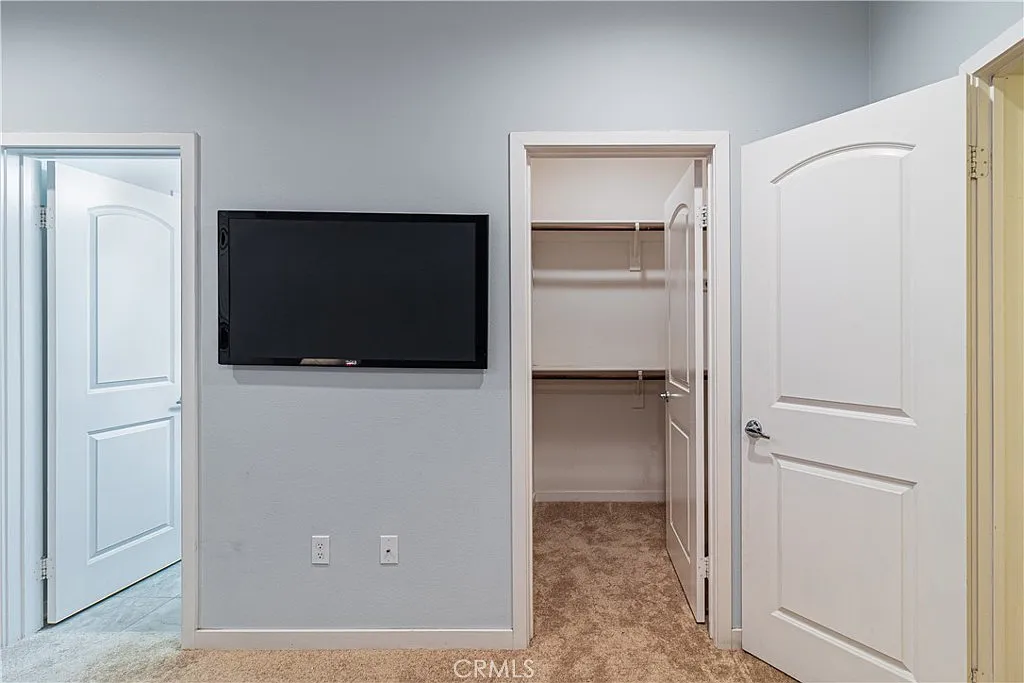 Room with wall-mounted TV, open doors to closet and another room, light gray walls, and carpeted floor.
