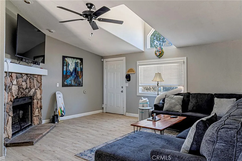 Living room with stone fireplace, mounted TV, gray sectional sofa, table, door, large windows, and wall artwork.
