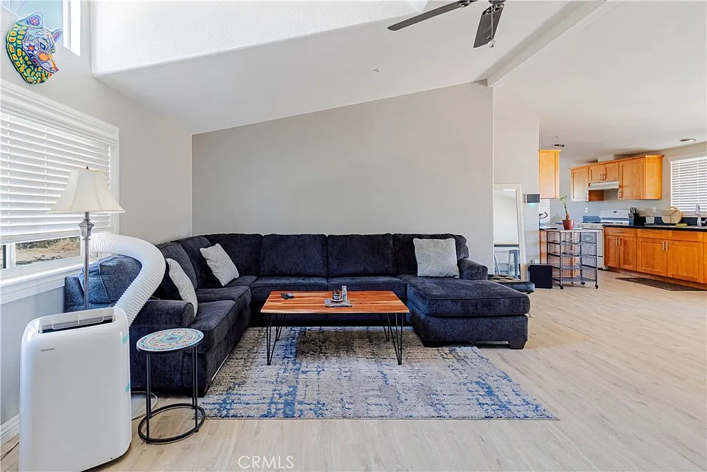 Living room with dark sectional sofa, coffee table, light rug, large windows, and open kitchen with wooden cabinets.