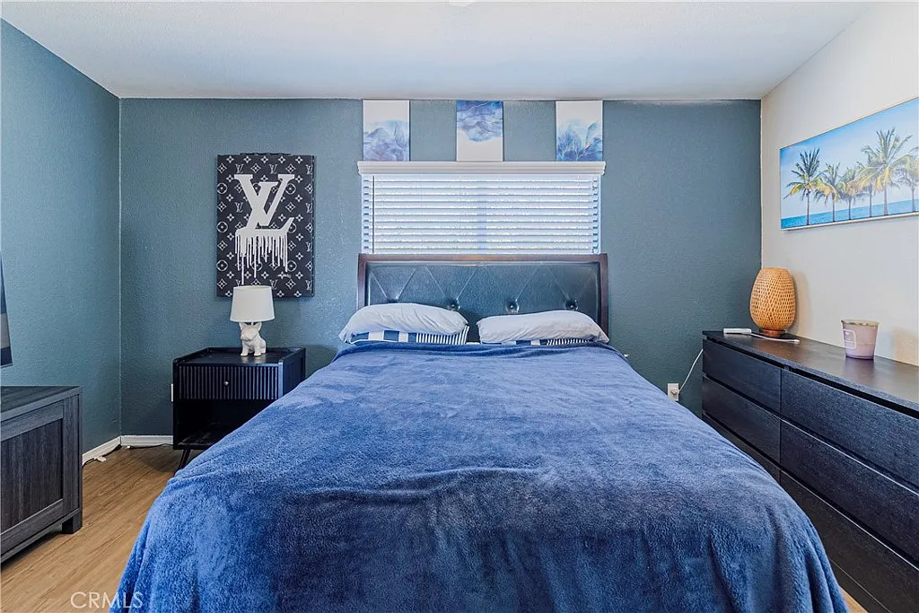 Bedroom with blue bedspread, headboard, decorative pillows, window, artwork, and wooden floor.