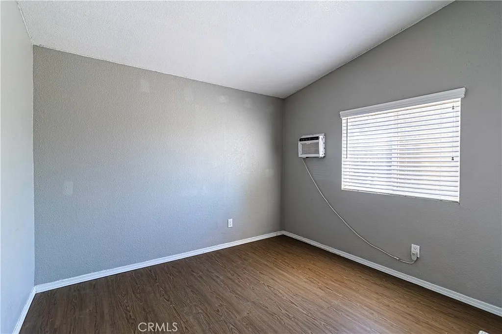 Empty room with gray walls, window with blinds, wooden floor, and wall air conditioner.