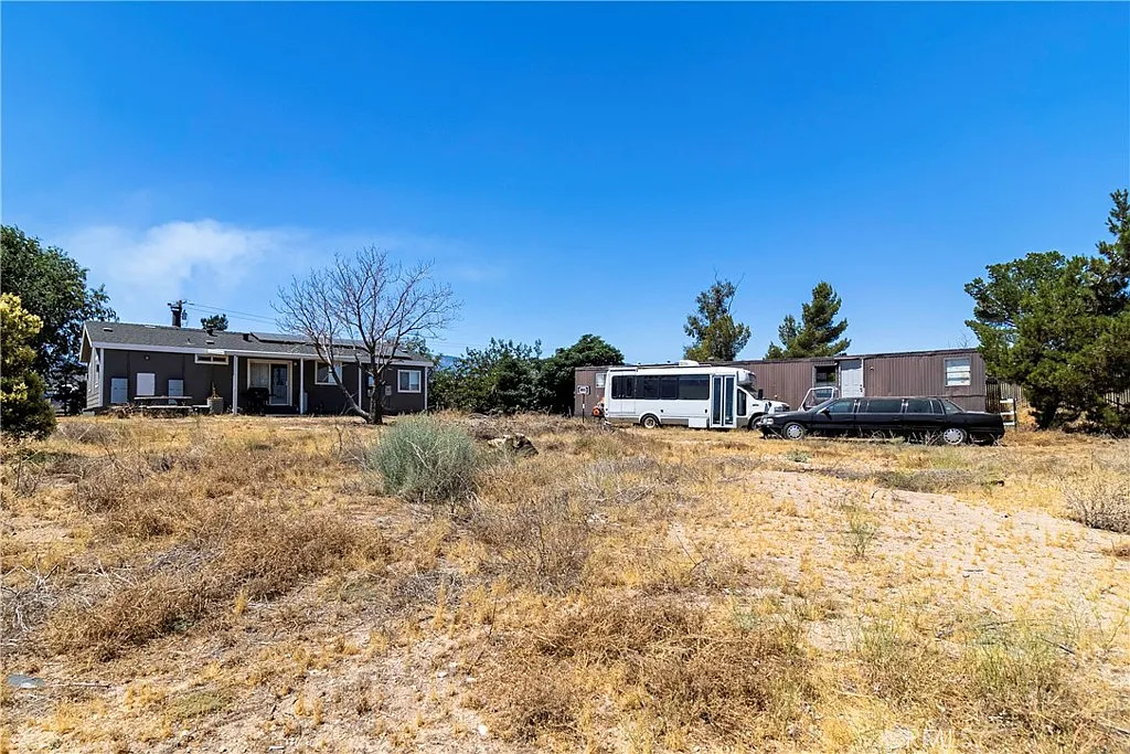 Property with house, bus, old car, dry grass, and sparse vegetation under blue sky.