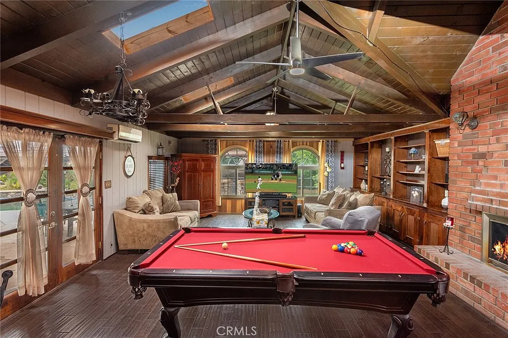 Game room with red pool table, wooden ceiling beams, fireplace, and large windows with green view