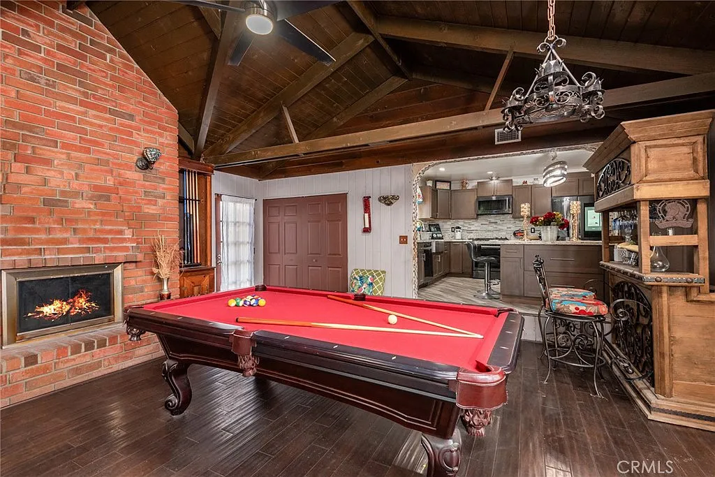 Game room with red billiard table, brick fireplace, wooden ceiling beams, kitchen view, and rustic cabinet with decorations