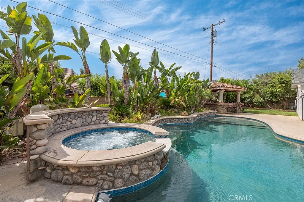Backyard pool area with stone hot tub, green plants, and gazebo