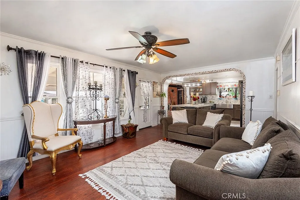 Living room with brown sofa, white pillows, patterned rug, ceiling fan, large windows, armchair, and view of kitchen
