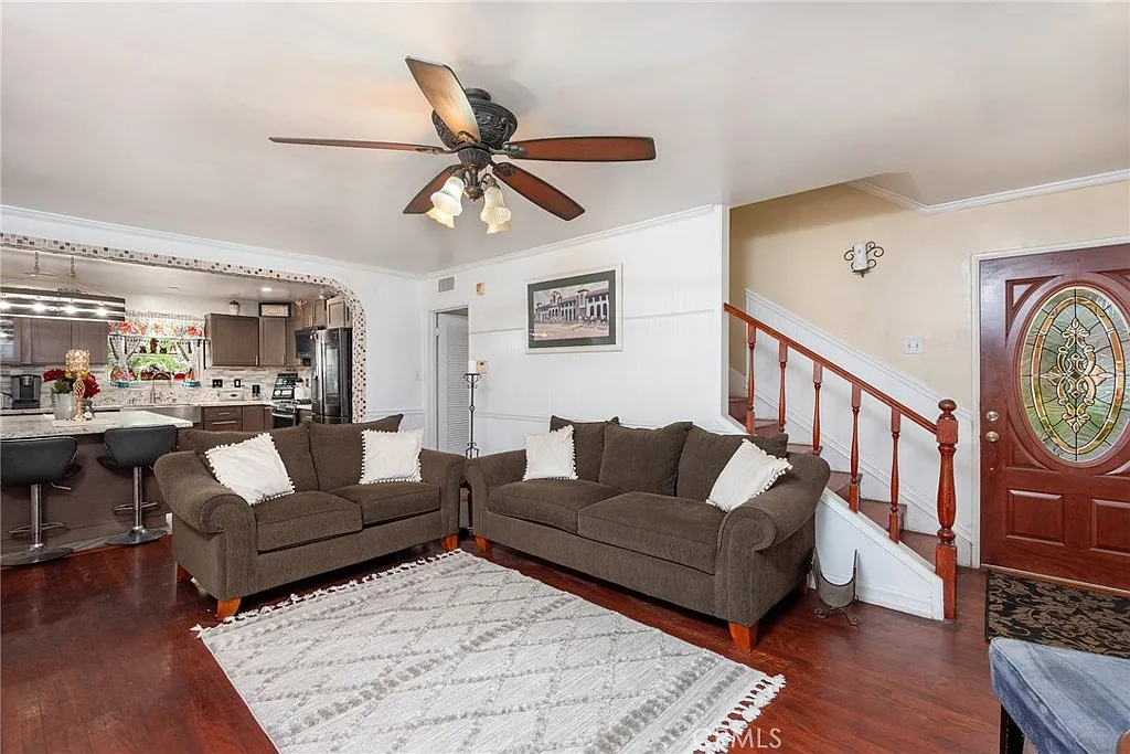 Living room with two brown sofas, ceiling fan, decorative rug, open kitchen view, and entrance door with intricate design