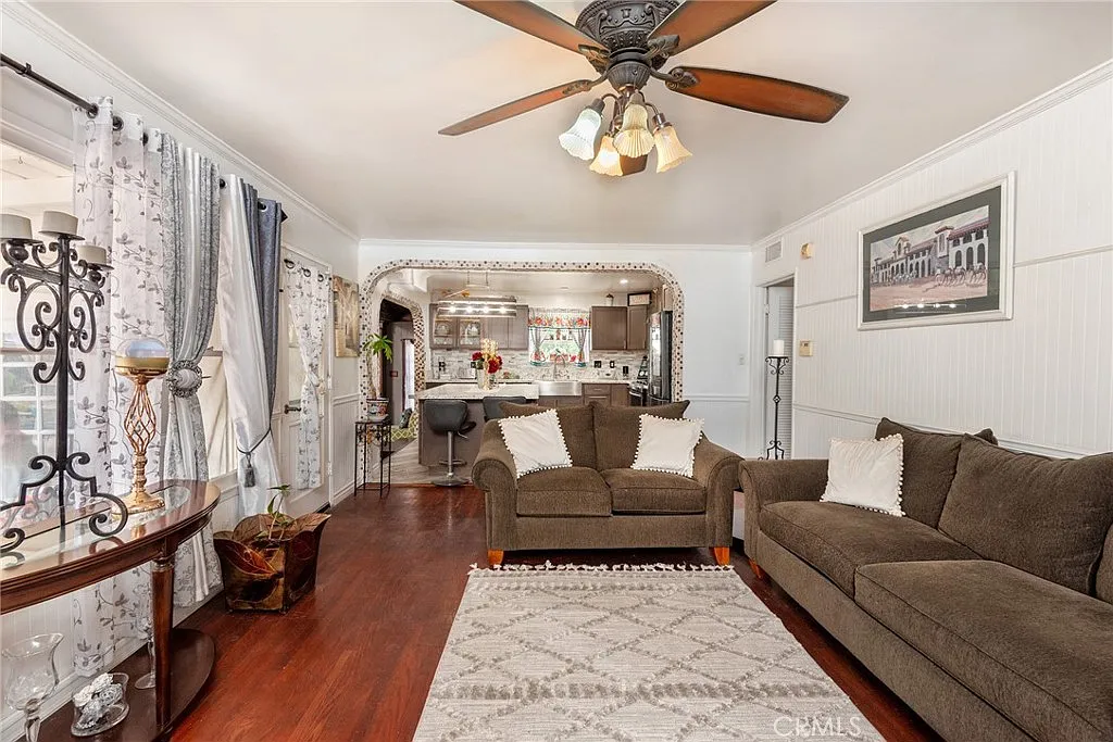 Living room with brown sectional sofa, wooden floors, curtains, ceiling fan, and kitchen with modern appliances in background