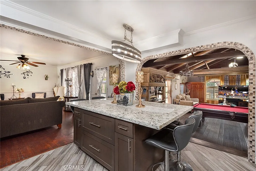 Kitchen island with granite countertop and bar stools, opening to living area with ceiling fan, pool table, archway, and colorful cabinets