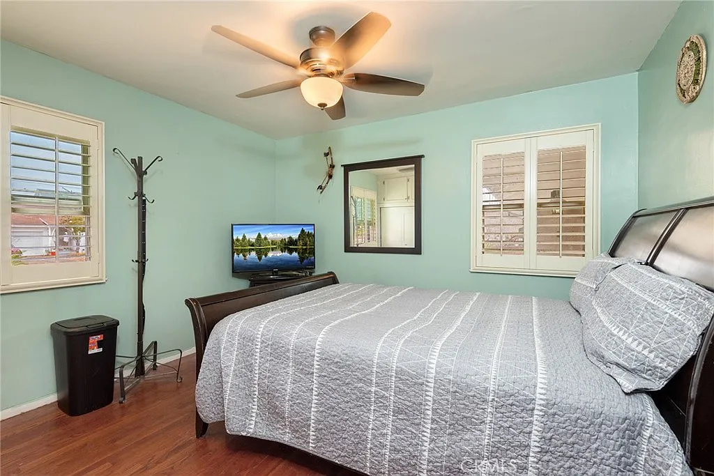 Simple bedroom with bed, gray duvet, ceiling fan, TV, light blue walls, coat rack, and small trash can