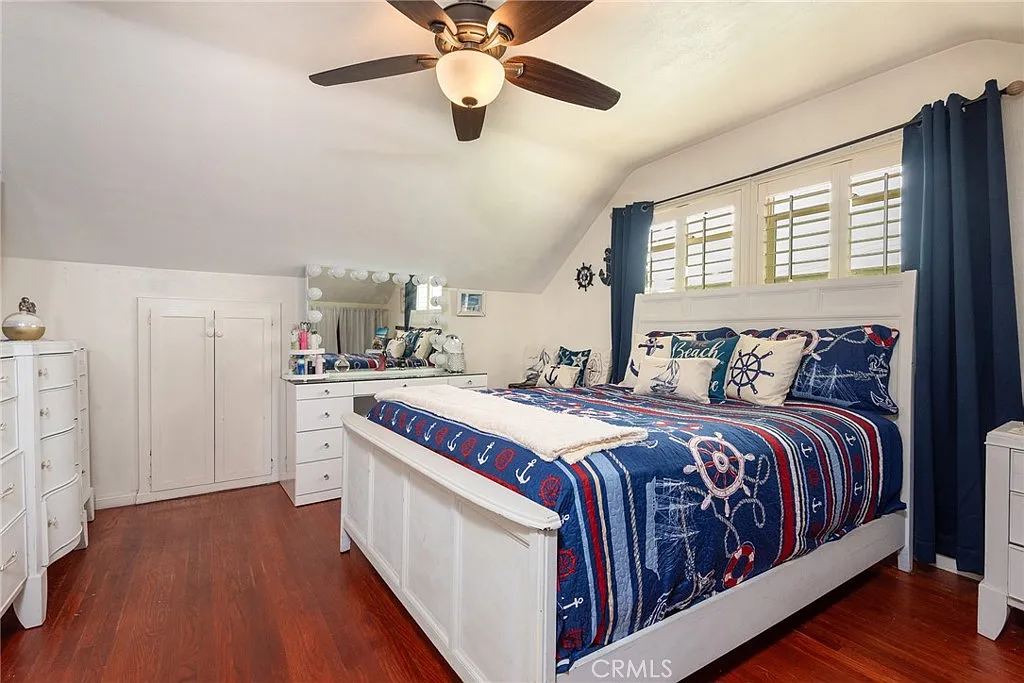 Bedroom with large bed, blue and white nautical duvet, ceiling fan, wooden floor, white furniture, and navy curtains