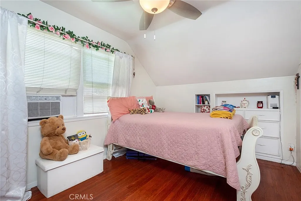 Bedroom with pink bedspread, ceiling fan, window, teddy bear on bench, and shelves with books
