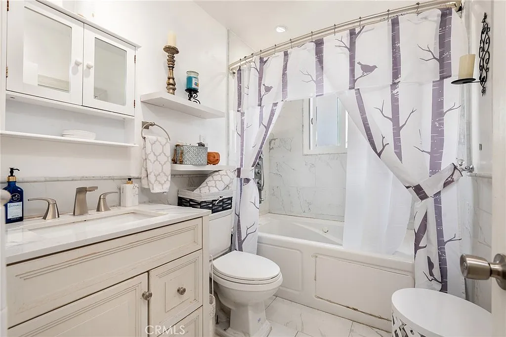 Small bathroom with white vanity, sink, toilet, bathtub with tree-design shower curtain, and decorative shelves