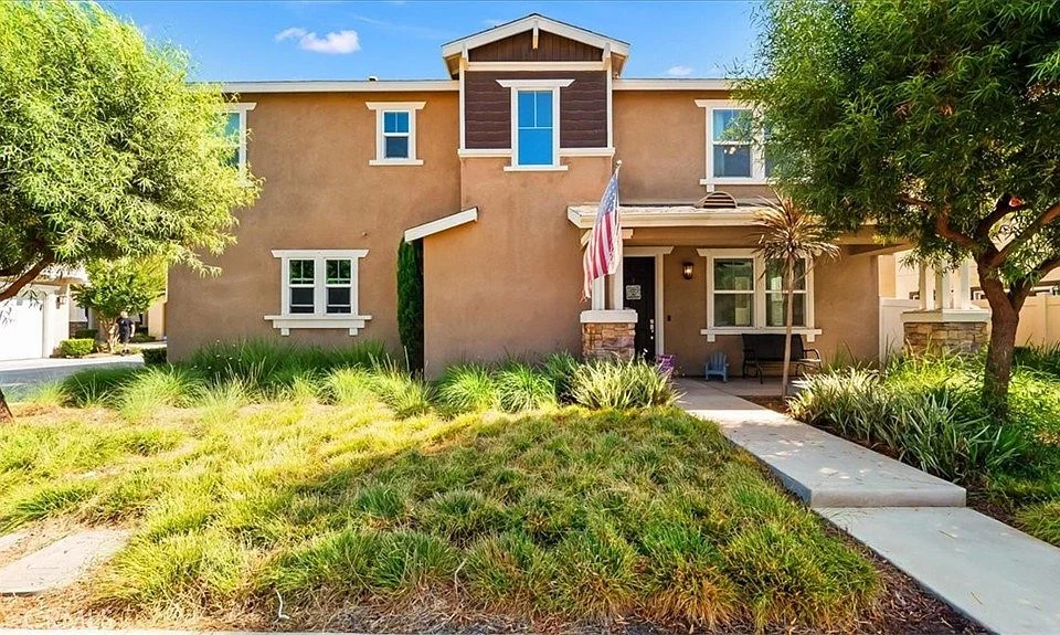 Two-story home real estate with brown exterior, porch, flag, grass, and shrubs.