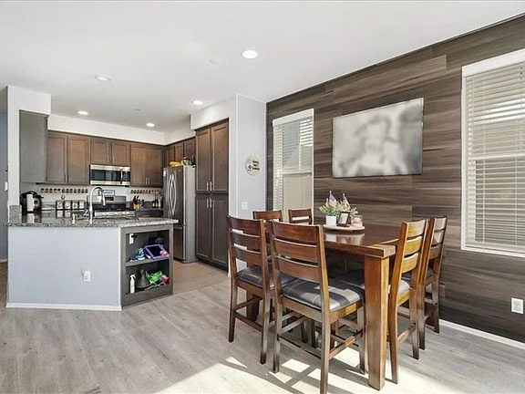 kitchen and dining area with wooden cabinets, stainless steel appliances, table set for six, light and wood-paneled walls, and large windows with natural light