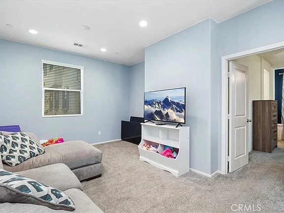 Light blue room with gray sectional sofa, television showing mountains, small white bookshelf, carpeted floor, window with blinds, and open doorway to another room