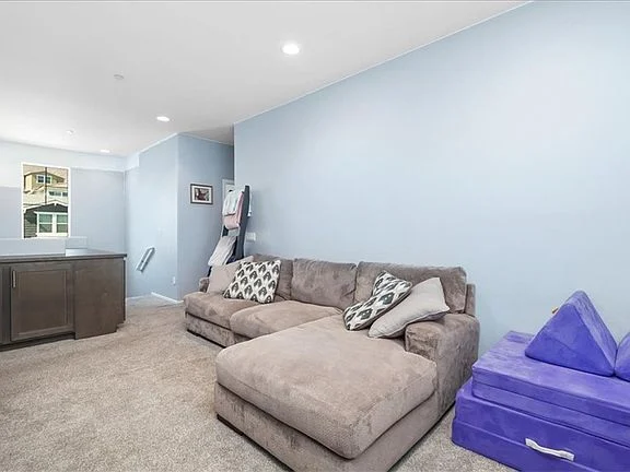 Living area with large gray sectional sofa, patterned pillows, light blue walls, carpeted floor, small table, and purple cushions