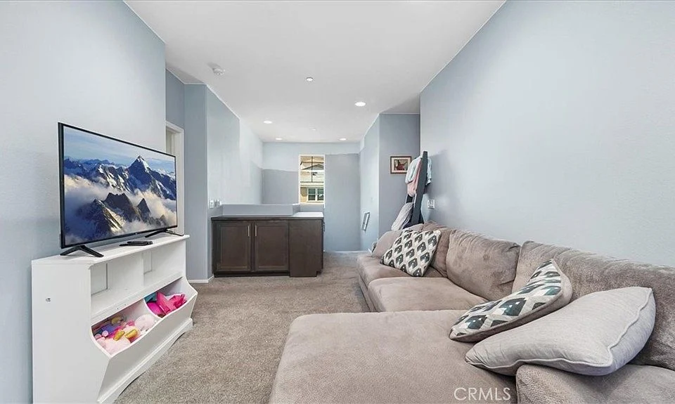 Living room with gray sofa, television showing mountain scenery, light blue wall, and open kitchen in background