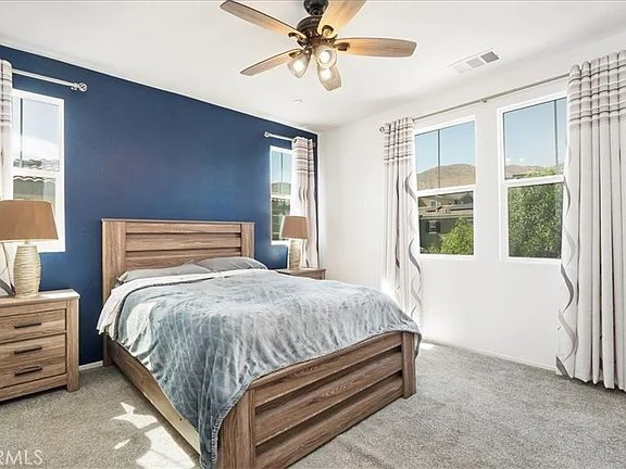 Bedroom with wooden bed, gray blanket, bedside tables with lamps, large windows with striped curtains, and blue accent wall