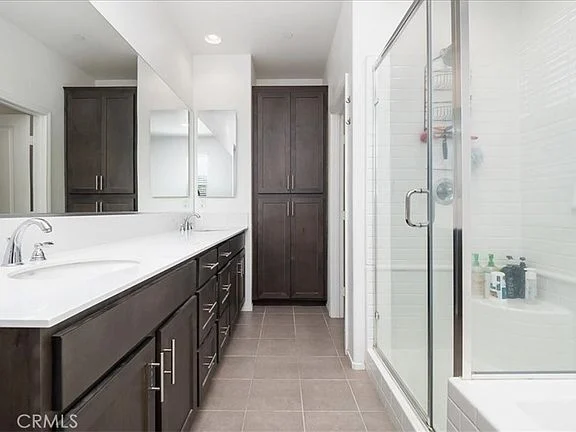 Bathroom with double vanity, dark wood cabinets, large mirror, glass shower, bathtub, and neutral tile floor