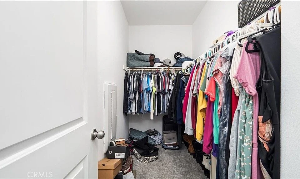 Closet with clothes hanging, boxes on floor, storage bins in corner, white walls, and visible door