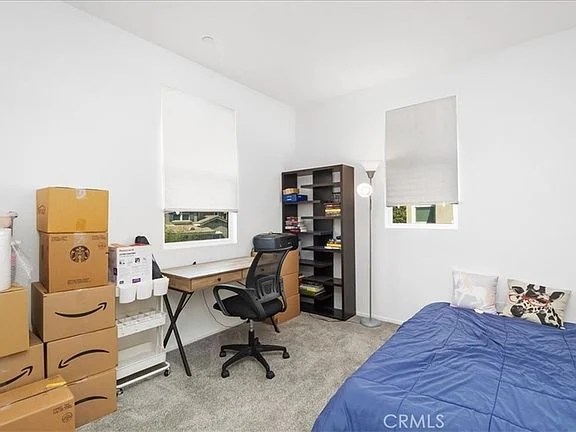 Bedroom with blue bedspread, desk with black chair, stacked boxes, bookshelf, light gray carpet, and two windows with white blinds