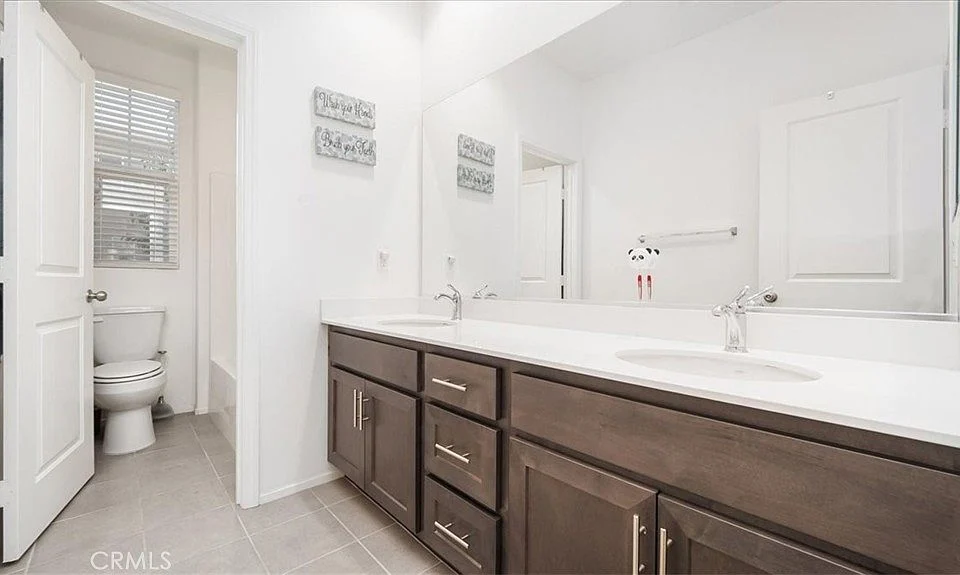 Bathroom with double sinks, large mirror, wooden cabinets, toilet in separate area, white walls, and tiled floor