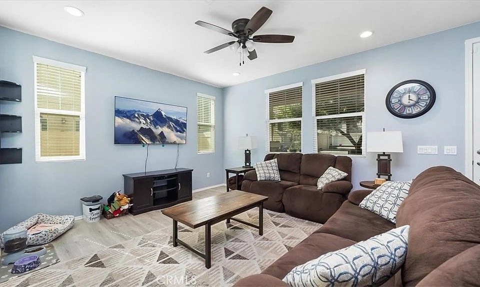 Living room with brown couch, wooden coffee table, wall-mounted TV, light blue walls, windows with blinds, and wall clock
