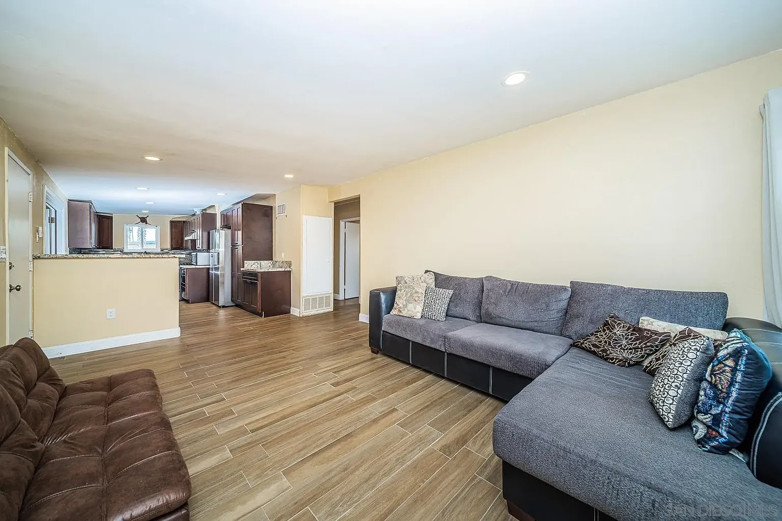 A living room with gray sectional sofa, brown couch, wood-like tile floors, and open layout to a kitchen.