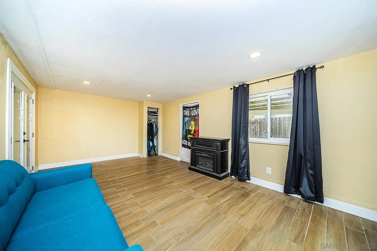 A light-colored room with wooden floors, blue couch, small black cabinet, window with dark curtains, and an open closet in the background.