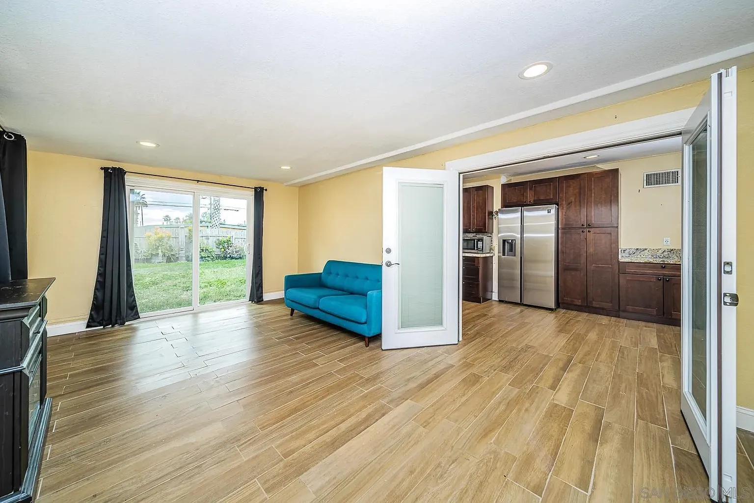 A bright living room with a blue couch, large windows, and open layout leading to a kitchen with dark wood cabinets and stainless steel appliances.