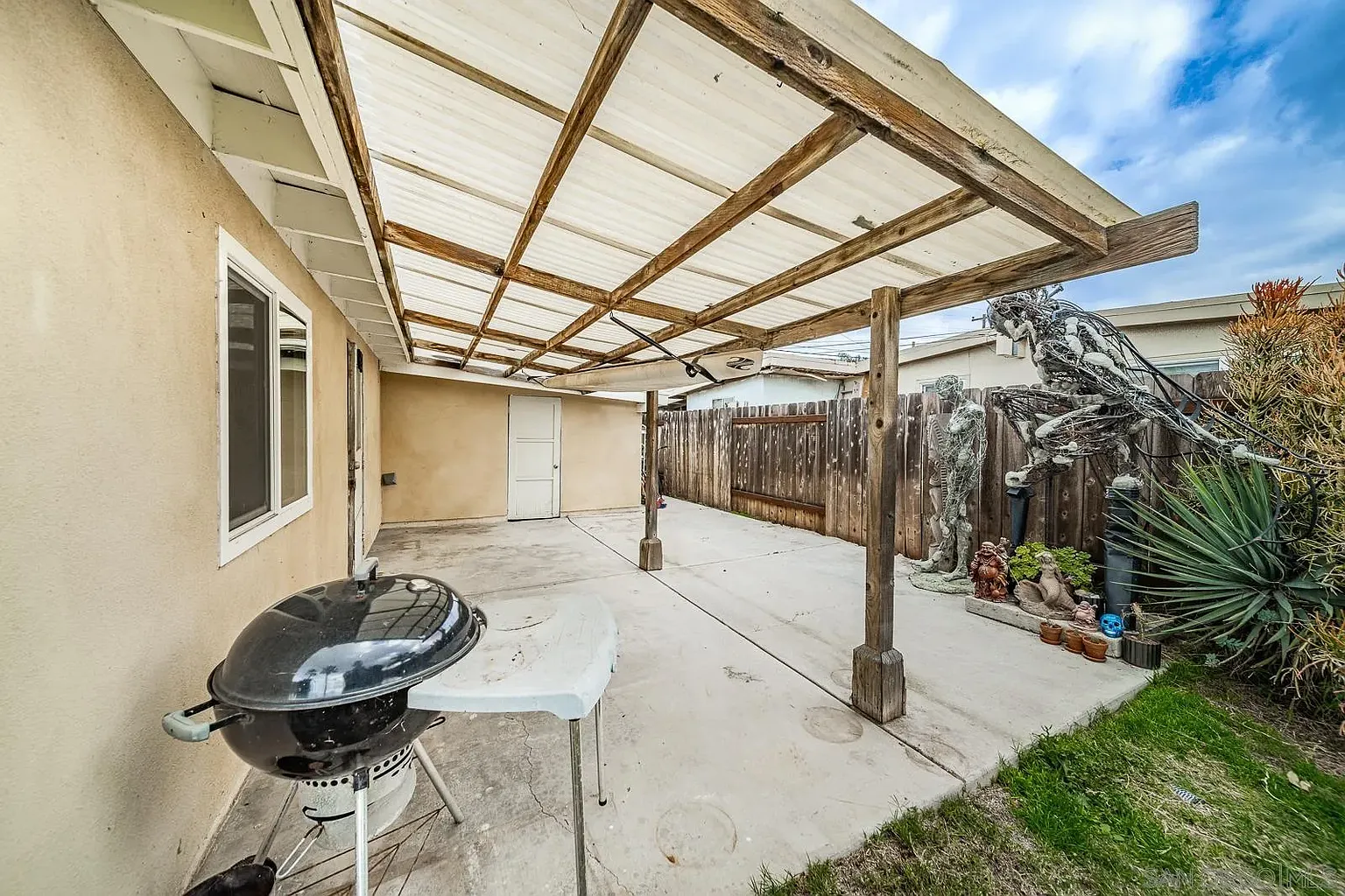 A patio with a barbecue grill, partially covered roof, wooden fence, and surrounding plants.
