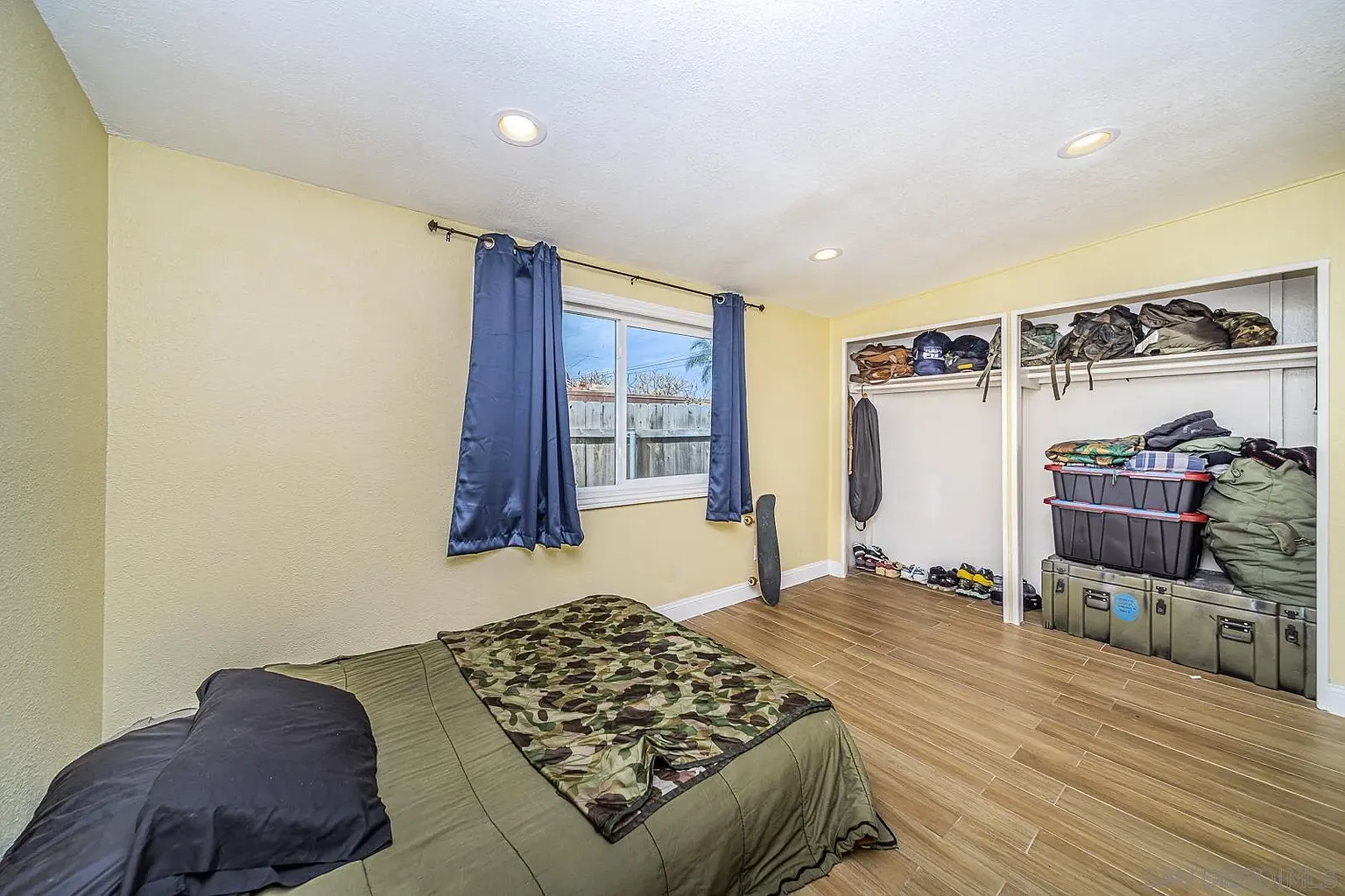 A bedroom with beige walls, large window with blue curtains, bed with camouflage bedding, and a closet with boxes and bags.