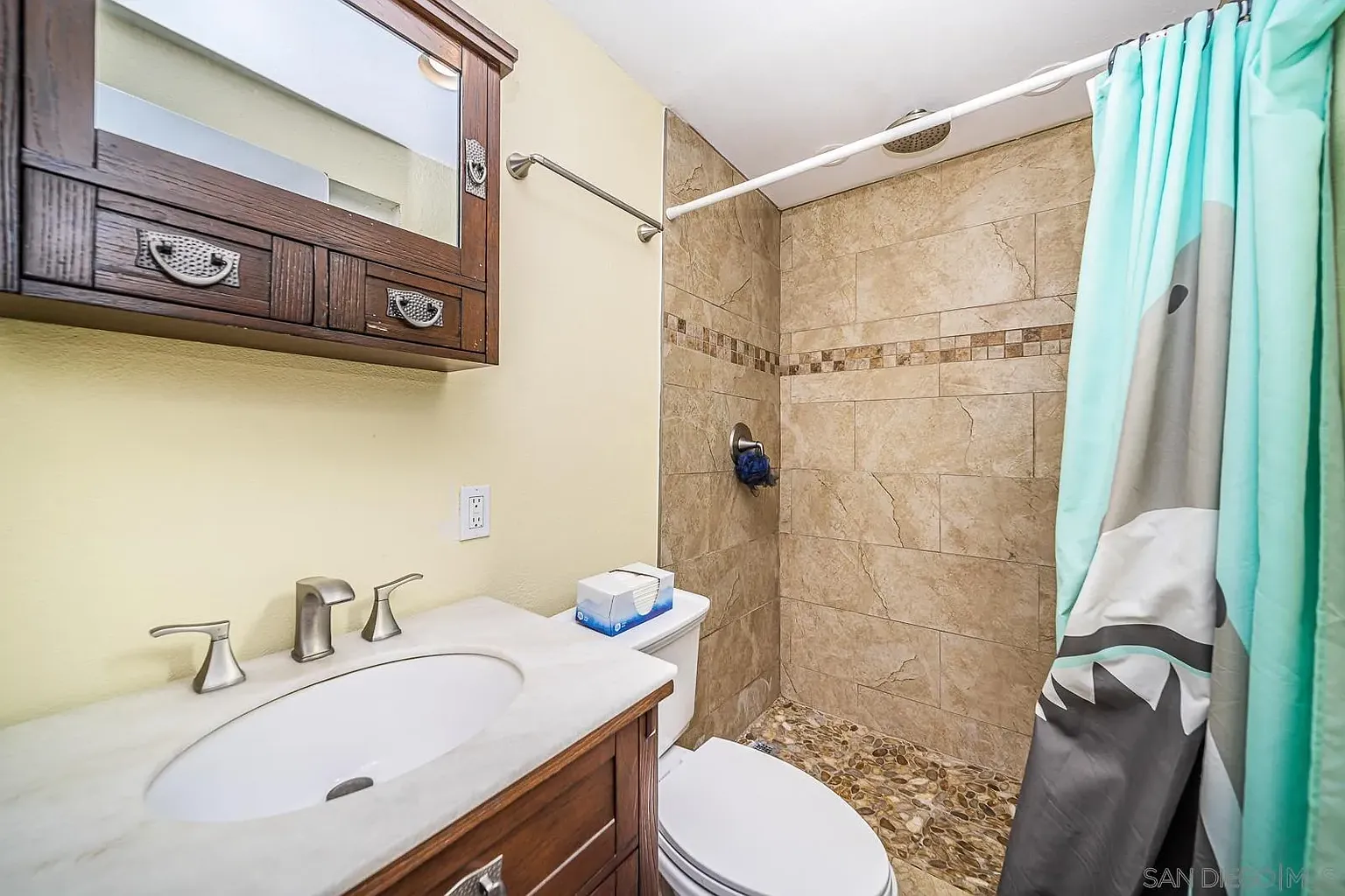 A bathroom with beige tile walls, glass shower, wooden cabinet with sink and mirror, toilet, and light blue shower curtain.
