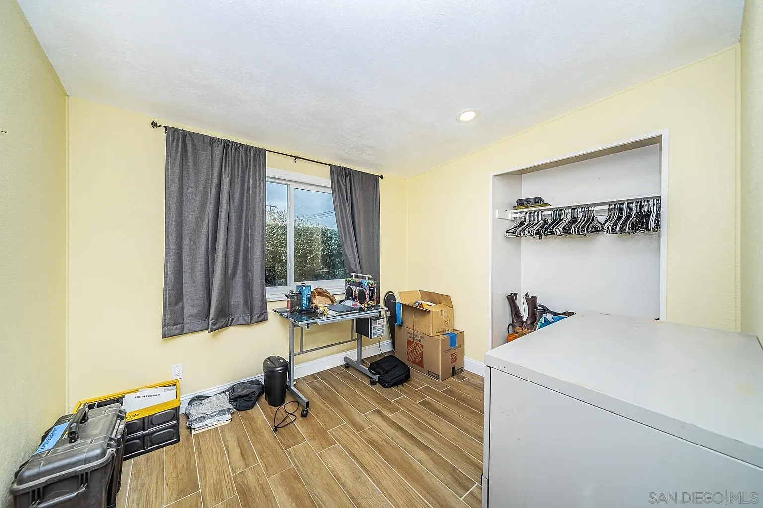 Room with yellow walls, window with gray curtains, closet with clothes, and boxes.