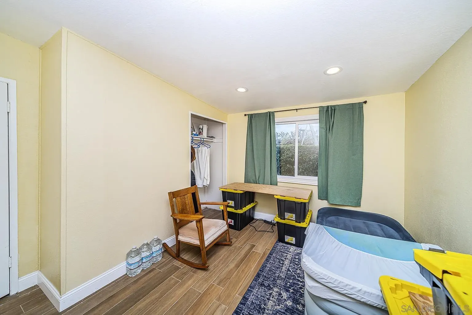 A small room with a rocking chair, desk with yellow storage bins, window with green curtains, closet, and bottled water on the floor.