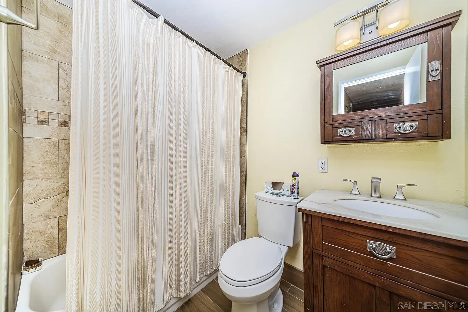 Bathroom with shower curtain, toilet, wood vanity, sink, mirror, and tile floor.