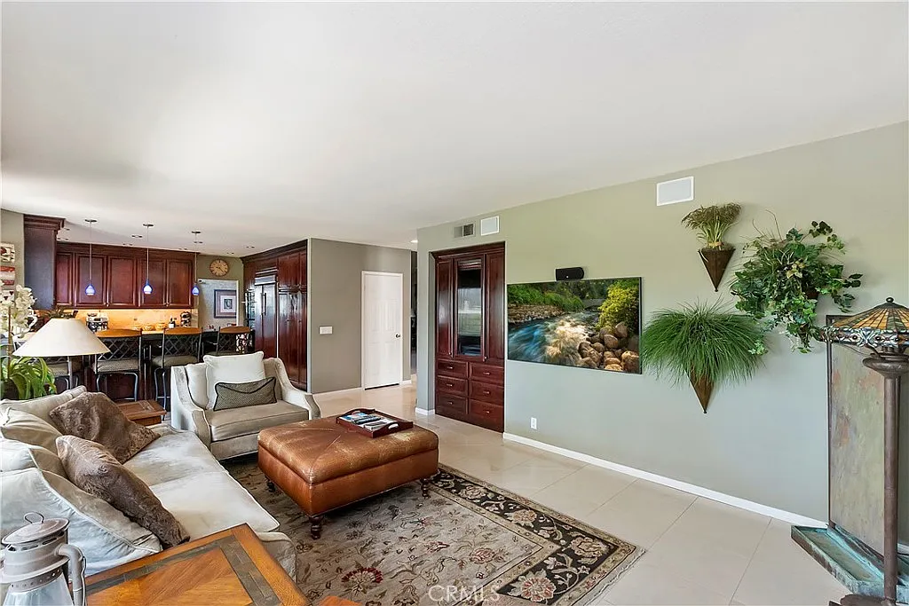 Living room with light sofa, ottoman, wall-mounted TV, plants, and patterned rug, with kitchen in background