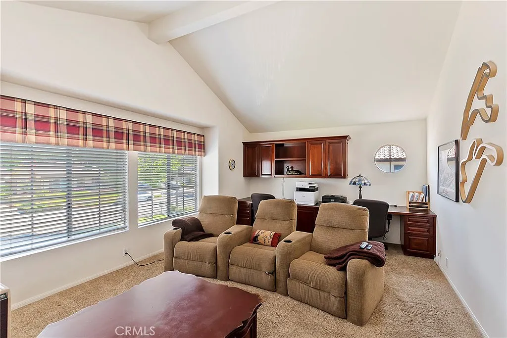 living room with high ceiling, beige recliners, wooden coffee table, window with plaid curtains, small desk near kitchen, light carpet, and white walls