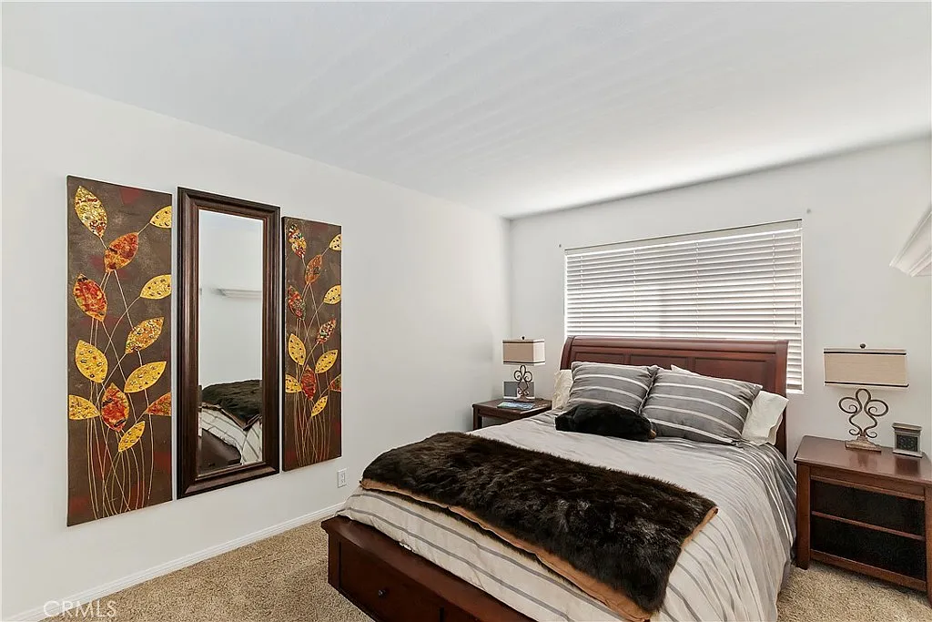 bedroom with bed in neutral colors, decorative pillows, fur throw, bedside tables with lamps, wall mirror, autumn leaf artwork, window with blinds, and carpeted floor