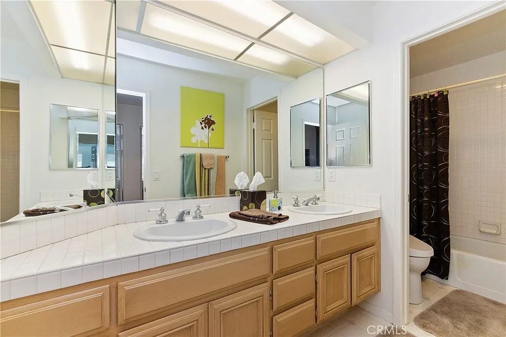 Bathroom with double vanity, mirrors, light wood cabinets, bathtub, green wall art, towels, and dark curtain near tub
