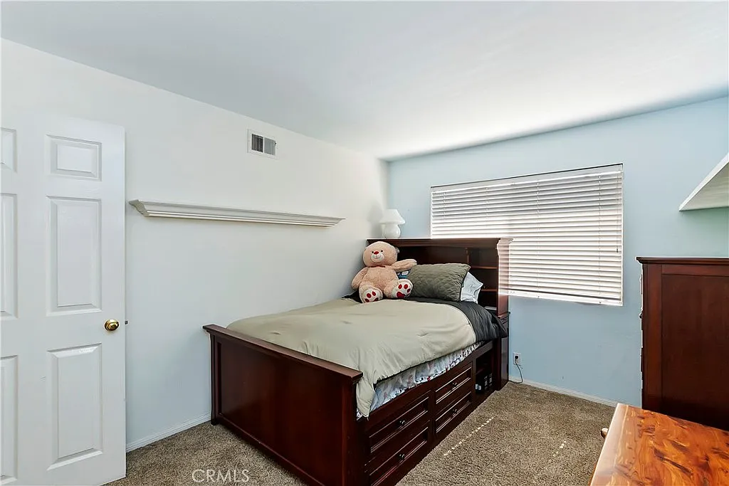 Bedroom with brown wooden bed with storage, gray and green bedding, large teddy bear, light blue walls, window with blinds, dresser, and shelf