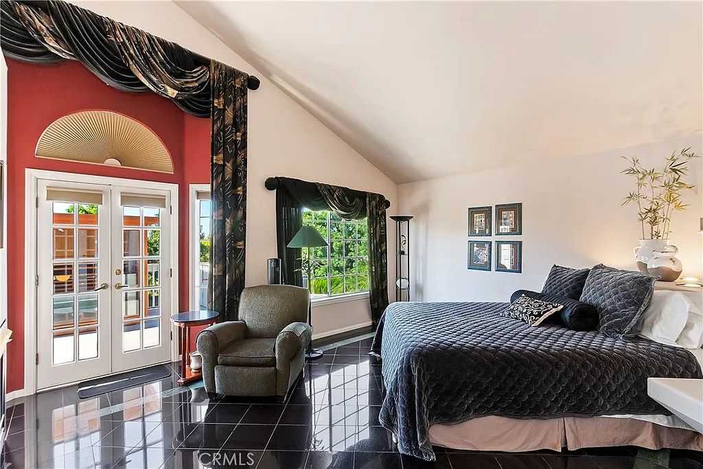 bedroom with large bed, dark bedding, green armchair, large windows with greenery view, high ceilings, and elegant drapes