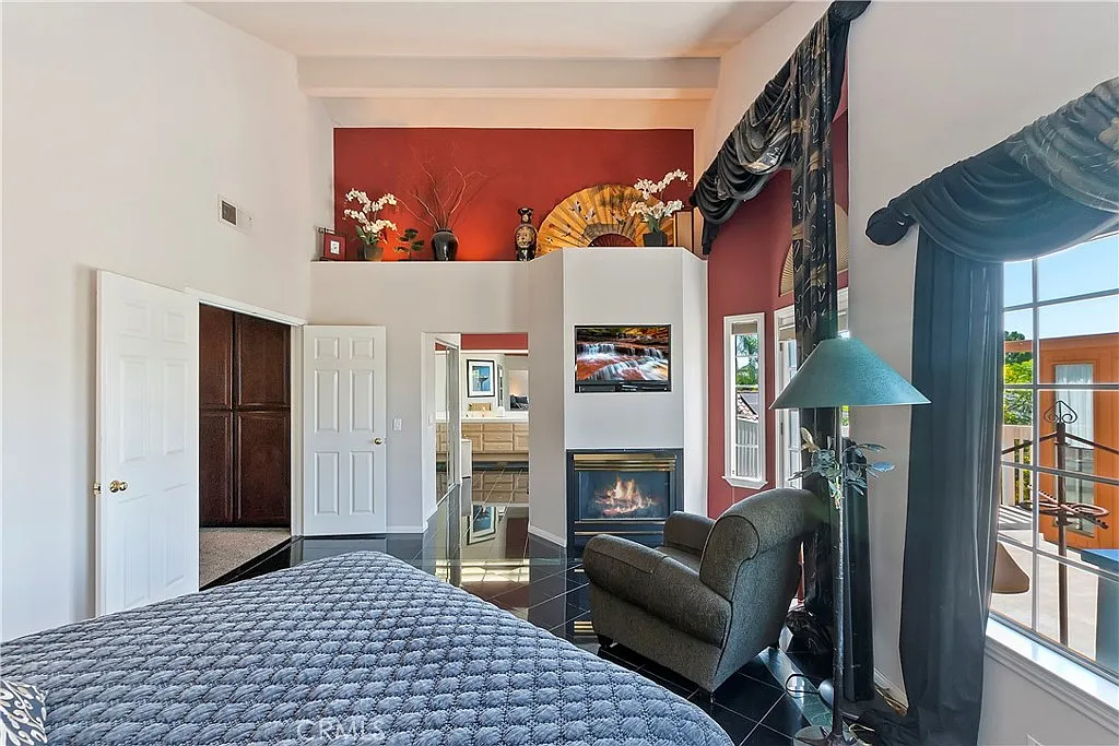 bedroom with large window and curtains, comfortable chair, fireplace, shelves with decorative items