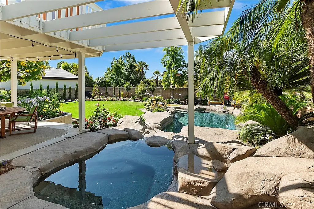 Backyard pool area with spa, landscaped gardens, trees, and white pergola patio